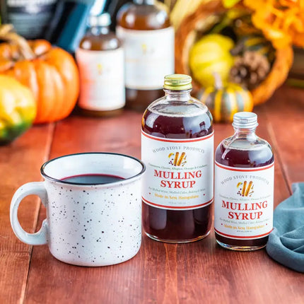 an autumnal tabletop covered with pumpkins and gourds and two bottles of mulling syrup alongside a mug of a tasty mulled beverage