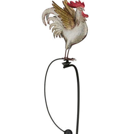 a farther photo of A metal garden stake featuring a colorful rooster with detailed feathers, perched on a rocker that moves in the wind.