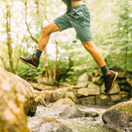 Person in green shorts and brown shoes jumping over a stream in a forest