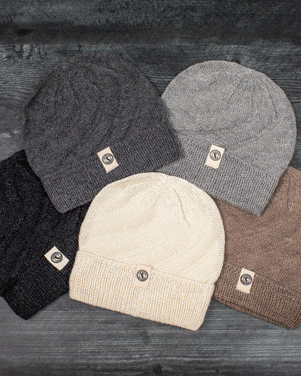 a grouping of neutral colored alpaca beanie style hats laid out on a gray wood floor - the hat colors include, from left to right, Charcoal, Gray, Cream, Silver, and Oatmeal