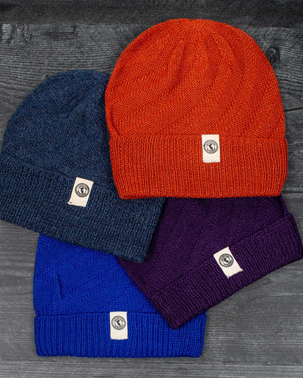 a grouping of colorful alpaca beanie style hats laid out on a gray wood floor. the hat colors include Denim, Rust, Blue, and Purple