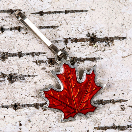 a handcrafted pewter and rust orange colored maple leaf zipper pull on a birch bark background