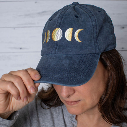 a model wearing the baseball cap, her head is titled downward showing off the embroidered details of the cap while gripping the brim of the hat with her thumb and forefinger