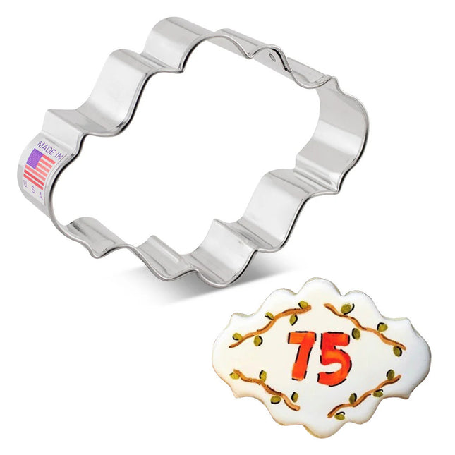 an oval cookie cutter with ornate edging beside a decorated cookie with lettering on it