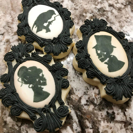 ornate edged oval cookies decorated with black icing to make a decorative frame around painted silhouettes of Victorian era skeletons painted in the style of a cameo