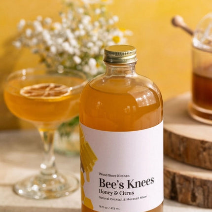 side view of Bee's Knees Mixer bottle on a tabletop next to a coupe style cocktail glass and a glass honey jar