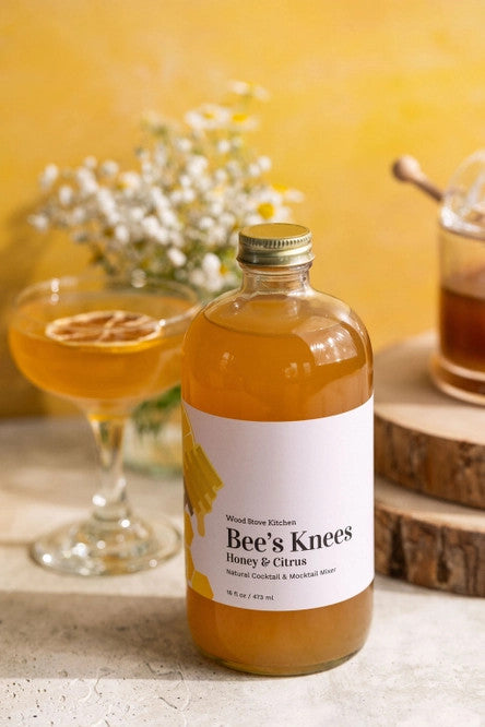 side view of Bee's Knees Mixer bottle on a tabletop next to a coupe style cocktail glass and a glass honey jar