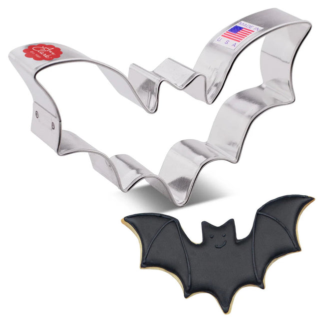 A tin cookie cutter in the shape of a flying bat with wings spread next to a fully decorated bat cookie with black icing, wing detail, and a cute smiling face
