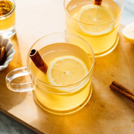 a tabletop with several glasses of warm amber liquid in clear rocks glasses and garnished with lemon slices and cinnamon stick