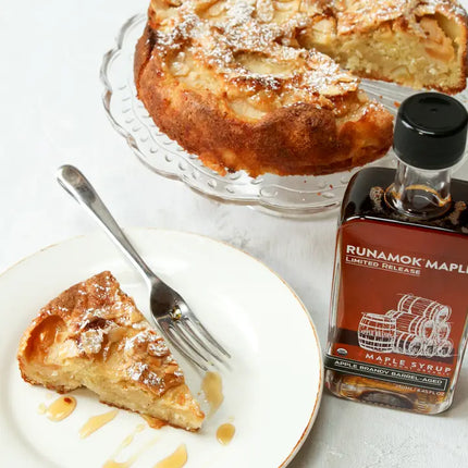 Slice of apple cake on a plate with a fork and a bottle of Runamok Maple syrup on a white surface.