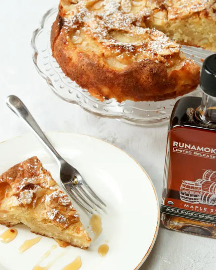 Slice of apple cake on a plate with a fork and a bottle of Runamok Maple syrup on a white surface.