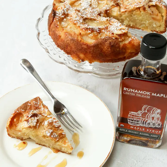 Slice of apple cake on a plate with a fork and a bottle of Runamok Maple syrup on a white surface.