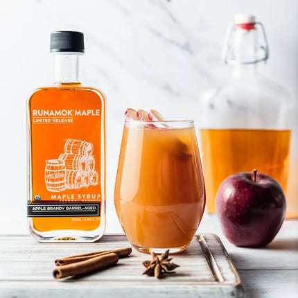 Runamok Maple syrup bottle with a glass of spiced apple cider, an apple, and cinnamon sticks on a white background.