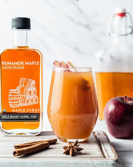 Runamok Maple syrup bottle with a glass of spiced apple cider, an apple, and cinnamon sticks on a white background.