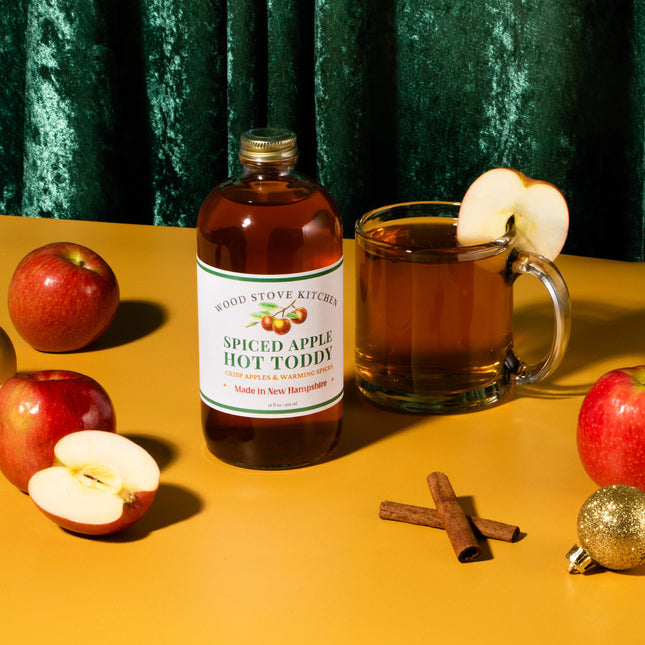 Spiced apple hot toddy bottle with a glass of hot toddy, apples, cinnamon sticks, and Christmas ornaments on a yellow surface.