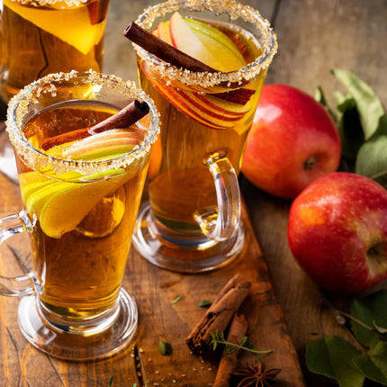 Two glasses of apple hot toddy with cinnamon sticks and apples on a wooden surface with apples and spices.