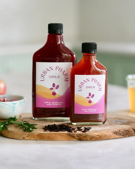 a bottle each of 200ml and 375ml Apple Rosemary Shrub on a wood board surrounded by fresh ingredients