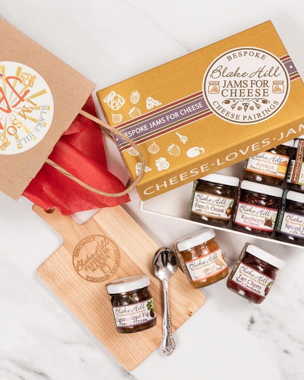 top down view of the cheese loves jam box on a marble countertop with the box partially open revealing jams in the box along with 3 jars outside of the box on a cheese board with a spoon