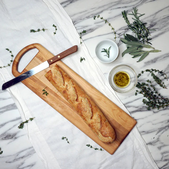 Baguette on a wooden cutting board with a knife, herbs, and olive oil on a marble surface.