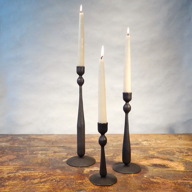 3 iron candlesticks of varied heights holding lit off-white taper candles sitting on a wood surface