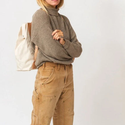 Woman wearing a brown sweater and beige pants with a white bag on a light background
