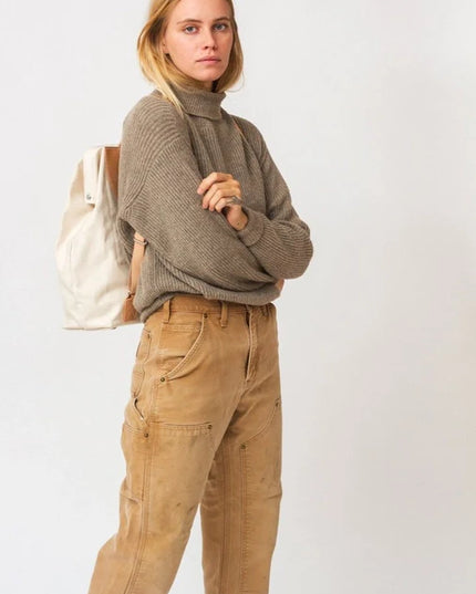 Woman wearing a brown sweater and beige pants with a white bag on a light background