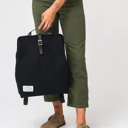 Person holding a black backpack wearing green pants and brown shoes on a white background