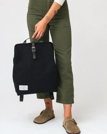 Person holding a black backpack wearing green pants and brown shoes on a white background