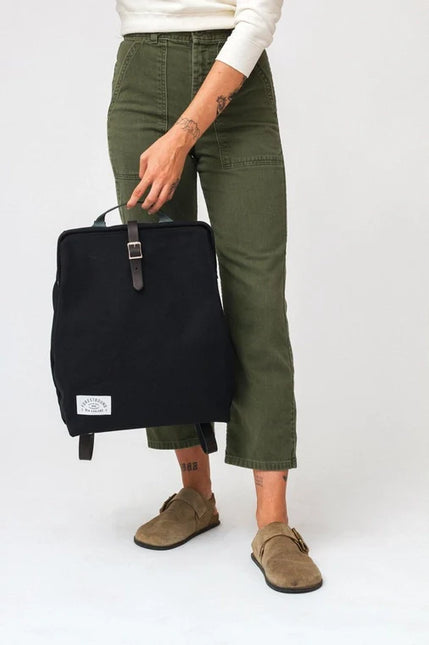 Person holding a black backpack wearing green pants and brown shoes on a white background