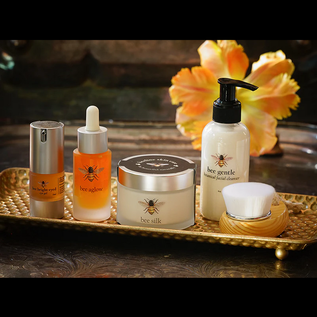 The Bee Renewed II skin care regimen line up of products, from left to right, Bee Bright-Eyed, Bee Aglow, Bee Silk Bee Gentle, and the facial brush set on a gold tray