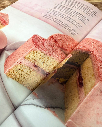 Heart-shaped pink and yellow cake on a marble surface with a book open above it.