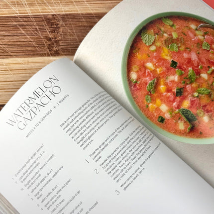 Open cookbook with watermelon gazpacho recipe and a bowl of watermelon gazpacho on a wooden surface.