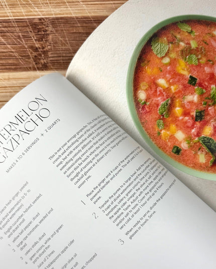 Open cookbook with watermelon gazpacho recipe and a bowl of watermelon gazpacho on a wooden surface.