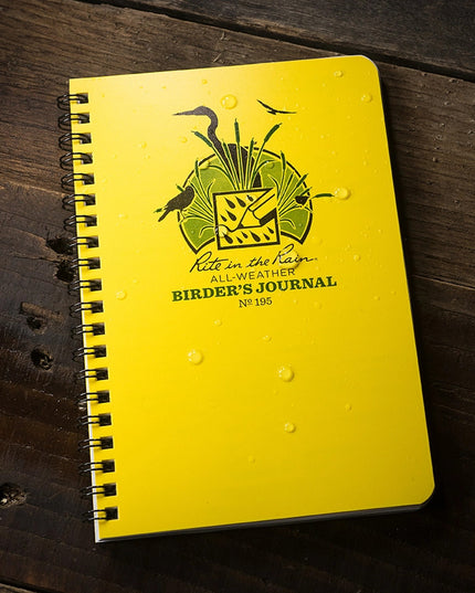 the weather-proof birder's journal with yellow cover laid on a dark wood surface with water beading on the cover