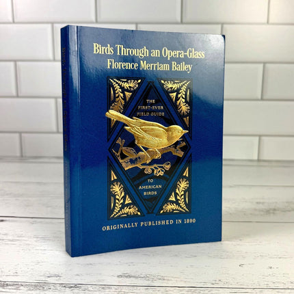 Blue book titled 'Birds Through an Opera-Glass' by Florence Merriam Bailey on a wooden surface with a tiled wall background.