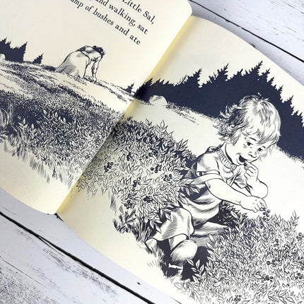 Illustrated book open to a page with a child sitting amongst low blueberry bushes