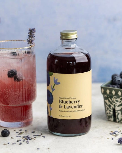 a tabletop scene with a bottle of Blueberry & Lavender mixer in the center and surrounded by glasses with brilliant purple beverages and a container of fresh blueberries