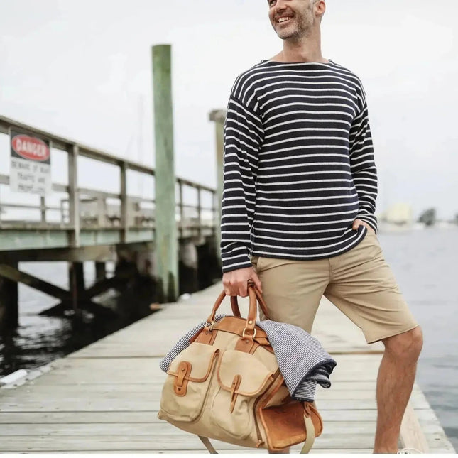 Man wearing a navy striped boatneck sweater holding a beige bag on a dock by water