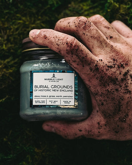 dirt covered hand holding a jar labeled 'Burial Grounds of Historic New England' against a grassy background