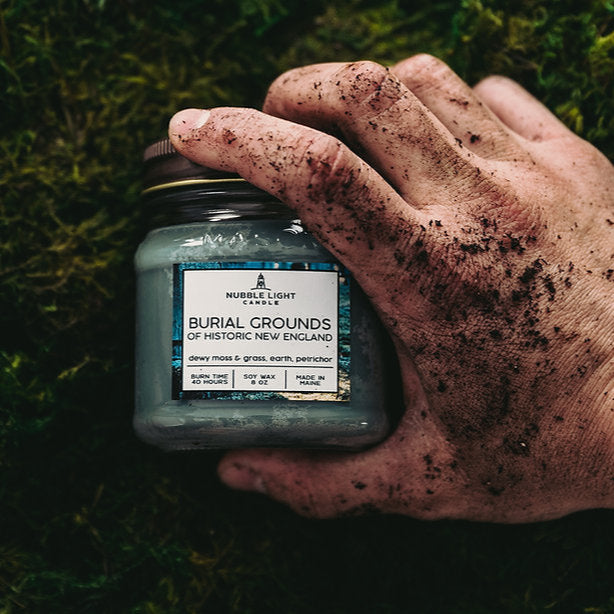 dirt covered hand holding a jar labeled 'Burial Grounds of Historic New England' against a grassy background