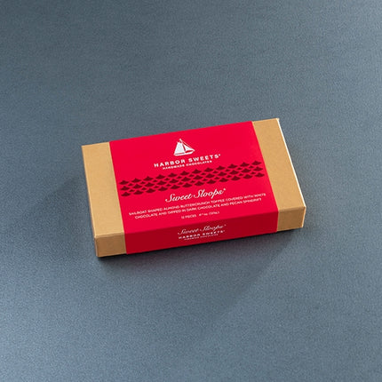 A rectangular box of Harbor Sweets' Sweet Slope chocolate, featuring a kraft brown cardboard body with a vibrant red paper band around the middle, set against a dark gray textured background.