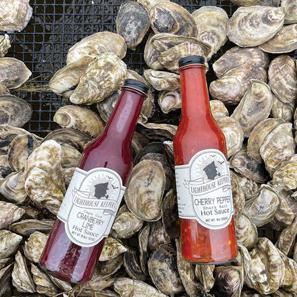 a bed of oyster shells with a bottle of cherry pepper hot sauce and a bottle of cranberry lime hot sauce laying on top