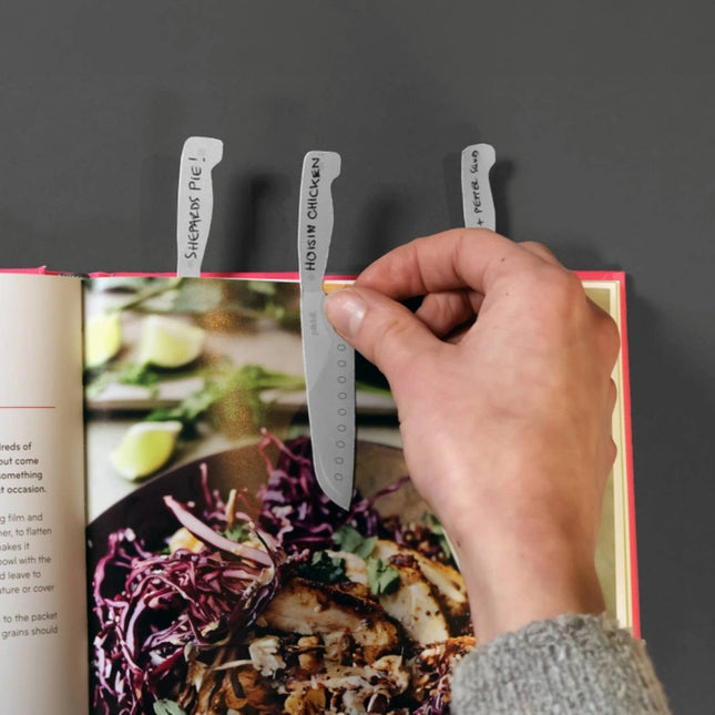 Hand using a silver bookmark with text over an open cookbook on a dark background