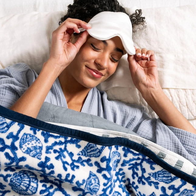 Woman wearing a sleep mask and robe, lying in bed with a blue and white seashell patterned blanket.