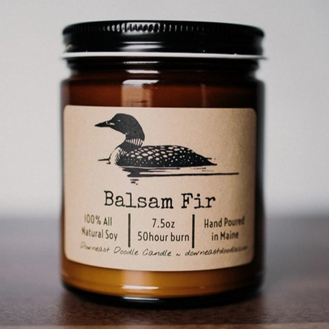 an amber glass candle with black metal top and kraft colored label with illustration of a loon above the text "balsam fir"
