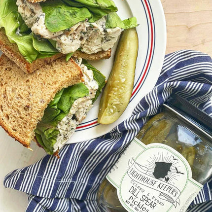 a tabletop spread with a chicken salad sandwhich on a plate with a pickle spear next to a jar of lighthouse keeper's pantry dill spear pickles