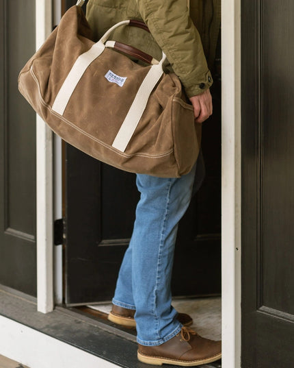 a model standing in a doorway with their brown waxed canvas duffle bag slung over one shoulder