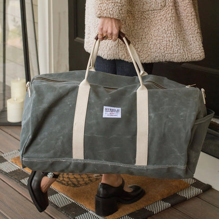 a close up of a model gripping the gray waxed canvas duffle bag by the leather handles