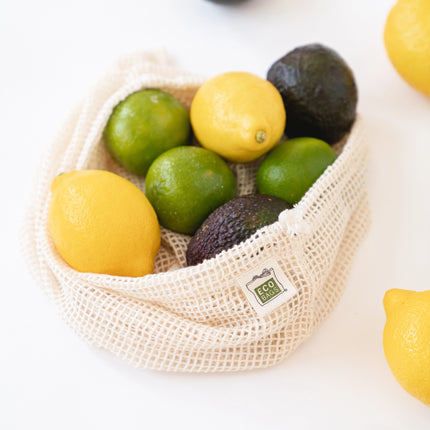 Fruit including lemons, limes, and avocados in a reusable mesh bag on a white surface.