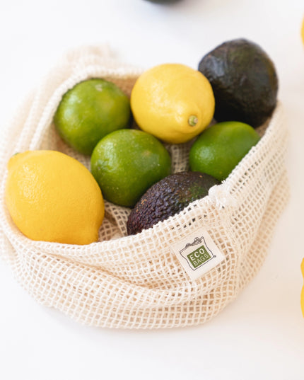 Fruit including lemons, limes, and avocados in a reusable mesh bag on a white surface.